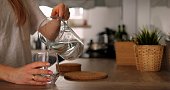 Faceless young woman pour pure water from jug to glass in kitchen. Anonymous girl in white t-shirt holds jar takes care of water balance