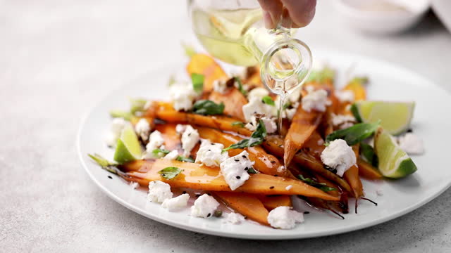 Pouring oil into a salad with roasted baked carrots and feta cheese, herbs on white plate, grey background. Vegetable salad with roasted young carrots, feta cheese.