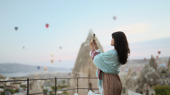 A young female tourist is taking photos of hot air ballons with her mobile smart phone from the balcony of the hotel during her travel.