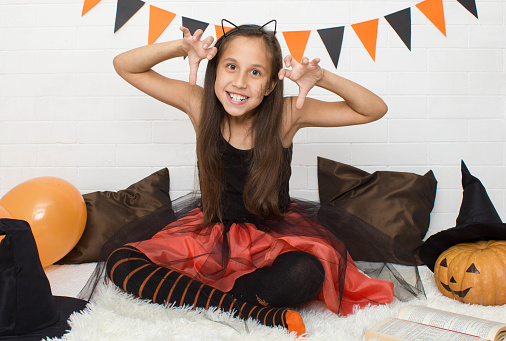 Funny blonde girl 7-8 years old in a witch costume for Halloween celebrates a holiday in a room decorated with flags, balloons and a pumpkin.