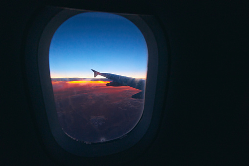 Beautiful sunrise view through the porthole during the flight. Plane wing above the cloudy colorful sunset