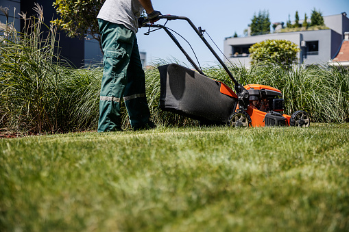 An unrecognizable man is maintaining a lawn with a mower in a public park.