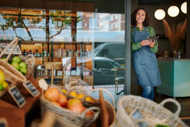 Portrait of a mature female delicatessen owner smiling while standing at the entrance to her shop with a digital tablet