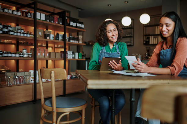 Smiling deli manager and an employee going over receipts at a table Mature female manager and a younger employee smiling while going over receipts at a table in an artisanal deli and cafe small business stock pictures, royalty-free photos & images