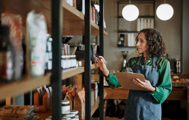 Mature deli owner taking inventory on store shelves using a clipboard Mature female delicatessen owner holding a clipboard while checking inventory of artisanal merchandise on some shelves in her shop small business stock pictures, royalty-free photos & images