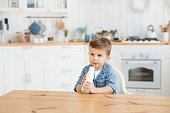 Cute baby toddler drinks milk on a blurred background of a light cozy kitchen. Handsome caucasian boy drinks vitamin rich organic lactose free yogurt with a straw. Copy space, product mock up. Tasty
