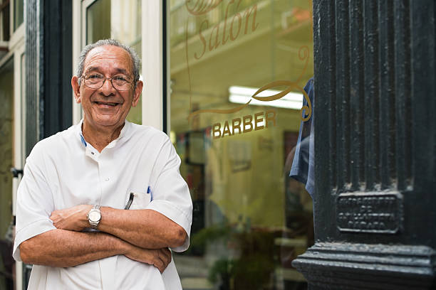 portrait of old barber smiling in hair salon senior hispanic barber in old fashion barber's shop, posing and looking at camera with arms crossed near shop window iberian ethnicity stock pictures, royalty-free photos & images