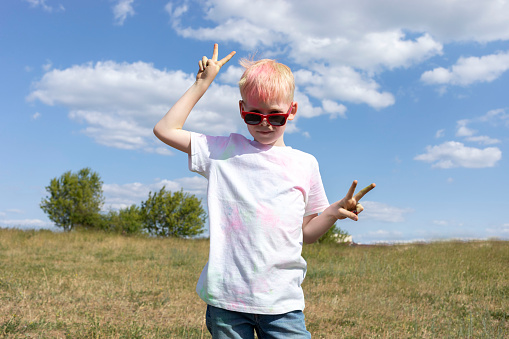 Little Blonde Boy With Colorful Paint, Powder On Clothes Posing On Holi Color Festival In Meadow. Emotional Happy Child. Blue Sky, Green Trees On Background, Sunny Day. Horizontal Plane