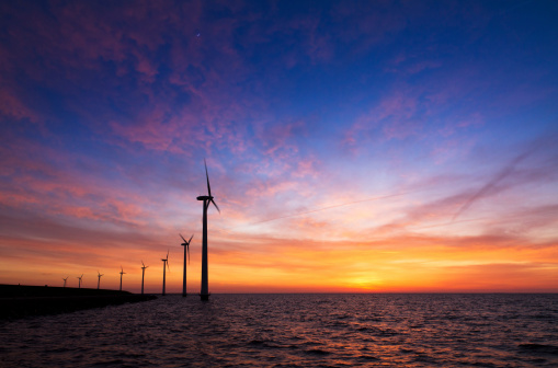 A large row of wind turbines along the coast at sunset with a small moon on top