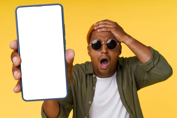 istock Surprised African man holds his head while showing phone with white blank screen on yellow background, concept for advertising. 1783742442