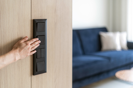 Selective focus on woman hand turning on or off black light switch on wooden wall at home. Power and energy saving. Modern living room interior design with furniture in blurred background