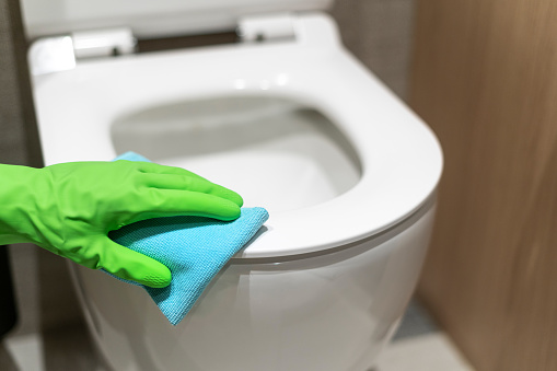 Cropped shot of female housewife hand in protective rubber glove hold wet cloth and wipe toilet seat at domestic restroom. Professional cleaner wash wc bowl at hotel. Cleaning service concept