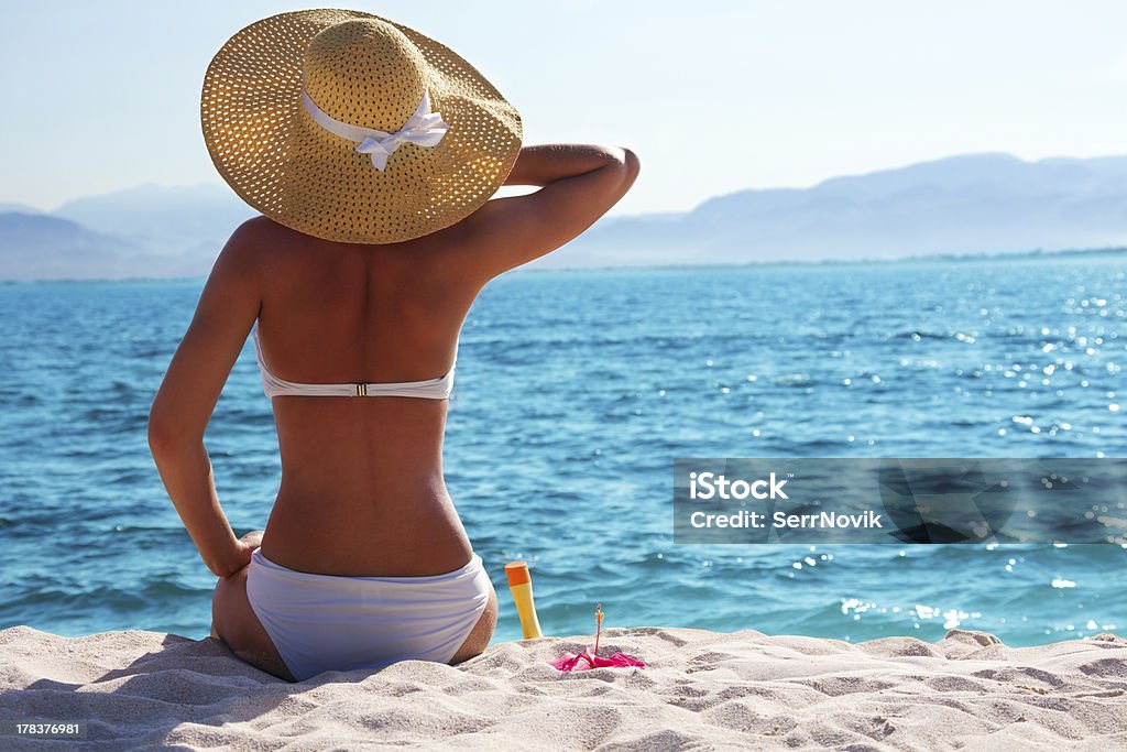 A woman with a sun hat is sitting on the beach woman in white bikini resting on the beach in straw hat Women Stock Photo