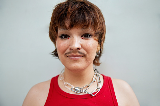 Headshot of individual in early 20s with short brown hair, mustache, earrings and necklace, standing against white wall, looking at camera with slight smile.