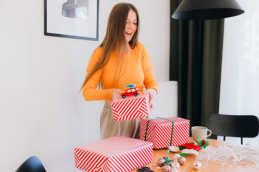 Smiling female with long hair in orange top enjoying winter weekend at home wrapping Christmas presents into the red boxes in modern living room during sunny day