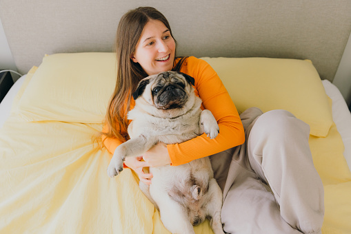 Happy woman with long hair in orange top relaxin on the yellow bed embracing with her cute and beautiful dog - pug breed
