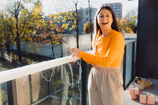 Smiling female with long hair in orange top enjoying winter weekend at home with her pug relaxing, decorating her balcony with Christmas lights and red gift boxes