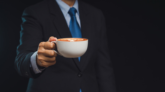 A businessman wearing a suit is holding a cup of coffee while standing against a black background Close-up photo