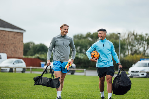 Two male football instructors heading to a pitch in the North East of England. They are carrying equipment talking together.