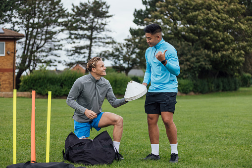 Two male football instructors heading to a pitch in the North East of England. They are carrying equipment talking together.