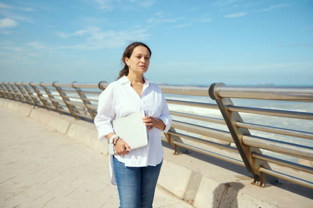 Young woman holding laptop, thoughtfully looking into the distance, strolling the street against seashore background