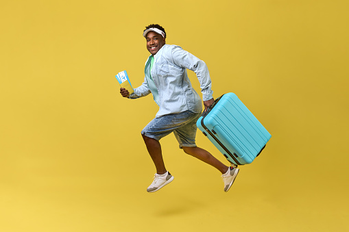 Happy African man in shorts and cap jumping with suitcase and tickets in hand