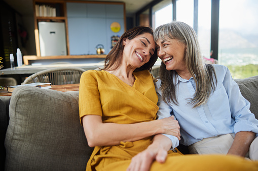 Senior woman and her adult daughter talking and laughing together while sitting on a living room sofa during a visit