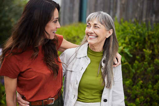 Senior woman and her adult daughter laughing together while standing arm in arm outside in a yard at home