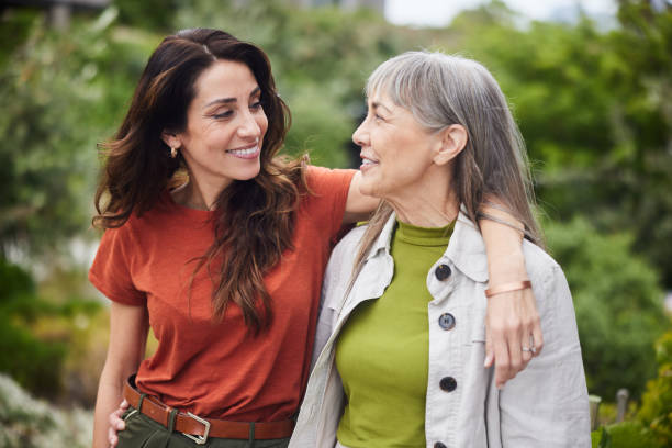 Smiling woman talking with her senior mom while walking outside Woman standing arm in arm with her senior mother and smiling while standing together outside in a yard at home adult age stock pictures, royalty-free photos & images