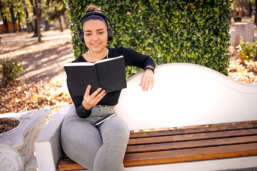 Beautiful woman enjoying in city park on sunny day in Autumn sitting on bench with book