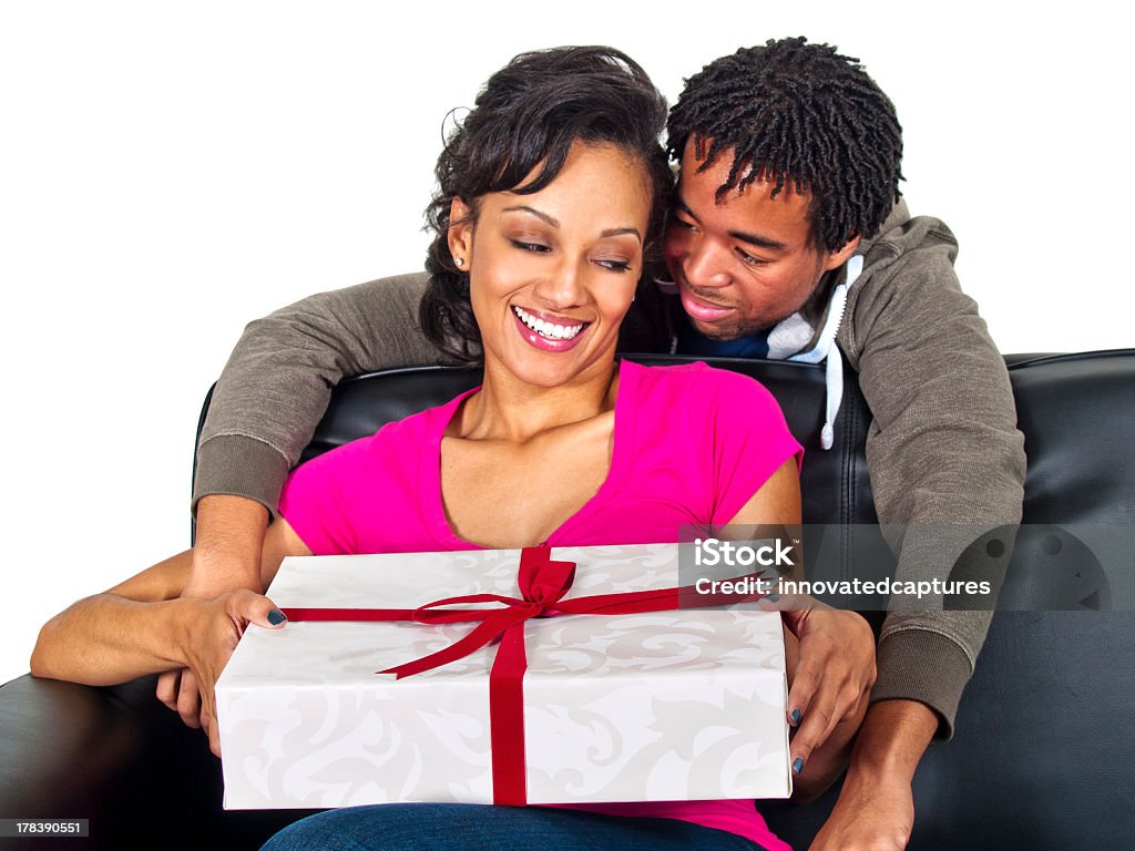 Black Male Giving a Surprise Gift to the Female Photo of a young African American couple sharing gifts.  The man is hiding a surprise for the female.  The young black couple is sitting on a couch. They are in a romantic relationship.  They are isolated on a white background. The image depicts a birthday or Valentines Day. Abundance Stock Photo