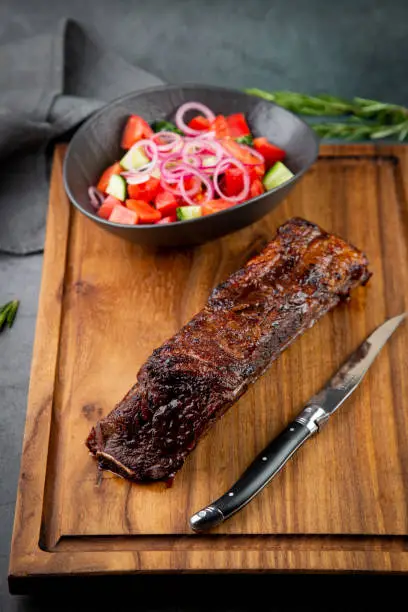 Photo of well-done steak with tomato and cucumber salad on a wooden tray, side view