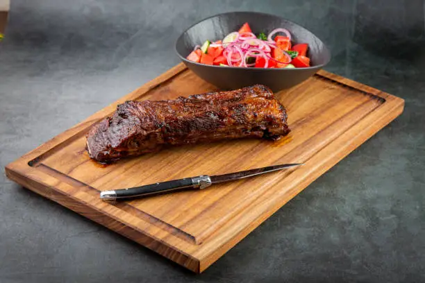 Photo of well-done steak with tomato and cucumber salad on a wooden tray, side view