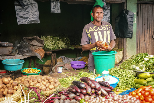 Ambatolampy, Madagascar - April 25, 2019: Typical street fruit and vegetable market at medium sized town. Unknown Malagasy shopkeeper putting carrots in green plastic basket
