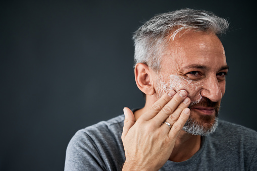 A grey-haired man putting on a moisturizer, taking care of his face skin.
