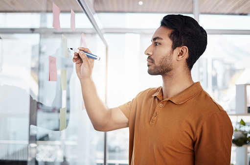 Writing, planning and focused man on glass for project management, moodboard and business priority planner. Asian person brainstorming ideas, job management and sticky notes for solution or reminder