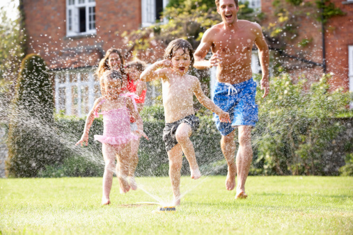 Family Running Through Garden Sprinkler Getting Wet Having Fun