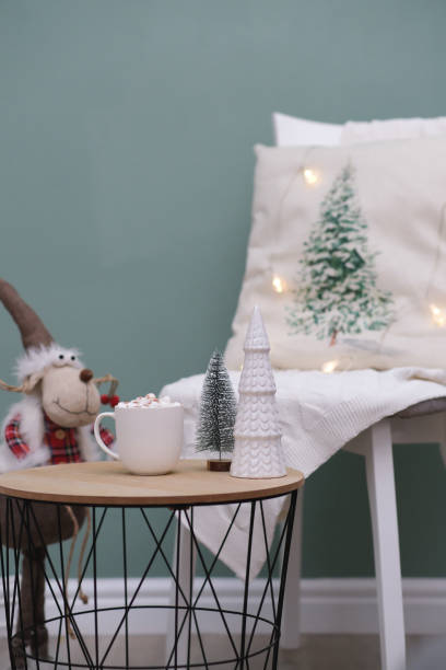 istock Christmas home decorations - cup of cocao with marshmellow, ceramic christmas tree on coffee table, christmas deer, char with pillow and blanket on green background. 1783951514