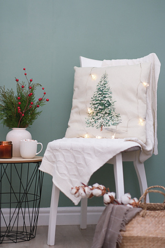 Christmas home decorations - chair with white blanket and pillow, burning candle, cup of cocao with marshmellow,fir branches, basket with cotton in vase on the coffee table on green background.