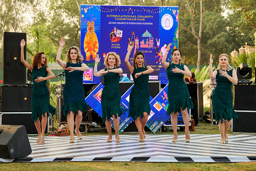 New Delhi, India - 10.12.2022 - Outdoor public park. Female dance group in green dresses dance on outdoor dancing floor at indian charity christmas fair, group of women dance expressively at outdoor stage