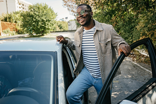 A young African american man opening car door with a smile on his face, while standing in in the parking lot