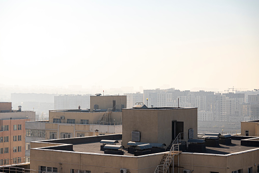 View of dark flat roofs with air conditioners, ventilation systems and waterproofing membranes of modern apartment buildings against the background of a city under construction shrouded in smog