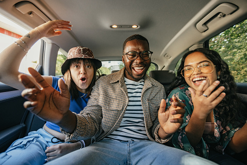 Group of three multiethnic friends having fun at the back seat of a vehicle while recording themselves with mobile phone