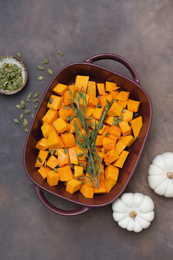 backed pumpkin in ceramic dish with oil, honey, seeds and rosemary with napkin on brown background. Top view.