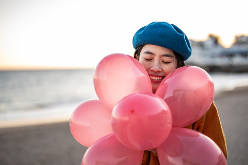 A cheerful young Chinese woman carries balloons in her hands, is by the sea and enjoys celebrating her birthday