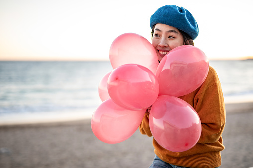 A cheerful young Chinese woman carries balloons in her hands, is by the sea and enjoys celebrating her birthday