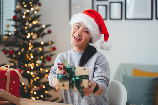 Asian woman sitting in home office decorated with a Christmas tree, smiles happily and sending Christmas present to camera.