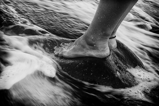 Black and white photograph of a person standing in the surf, with their feet partially submerged in the water
