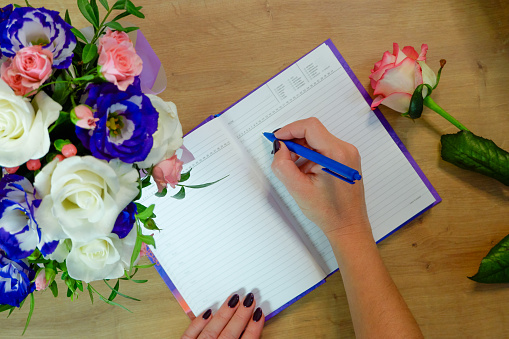 Female hands writing in open notebook and bouquet of roses on old wooden table. Planning notebook with pen, woman makes entry in her diary on table.