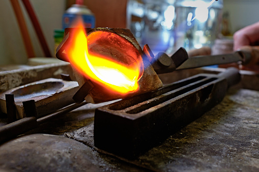 Heated to red-hot melting point by a blowtorch, pure silver being poured from a jeweler's crucible into a mold among tools of the trade.